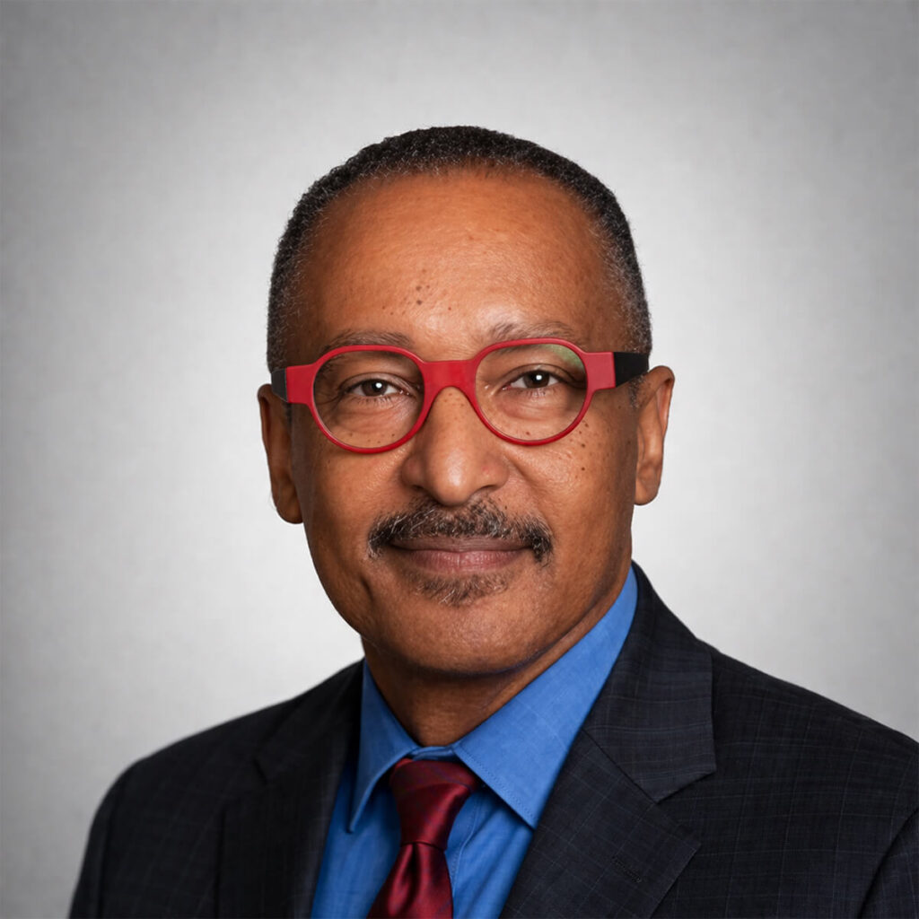 Professional headshot of a man wearing red round glasses, a blue shirt, and a dark suit with a red tie.