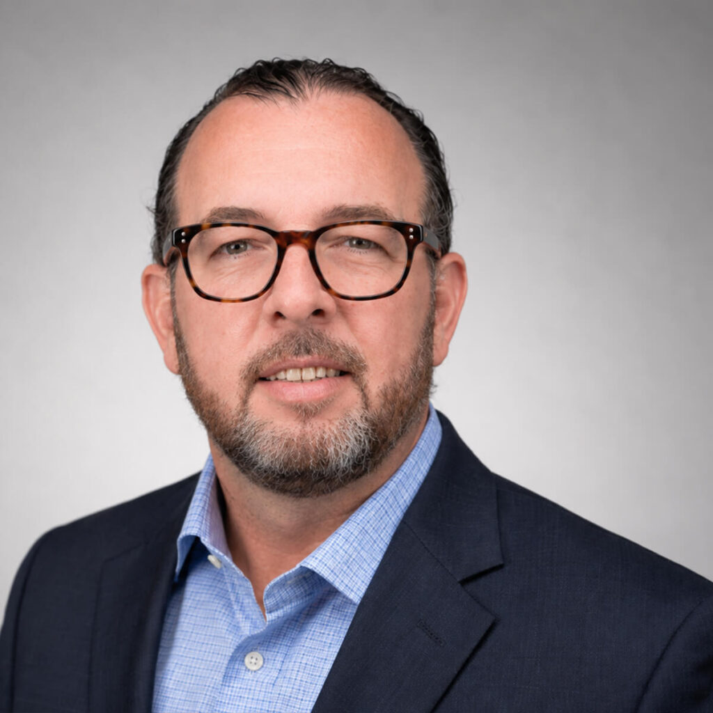 Professional headshot of a man wearing tortoiseshell glasses, a navy blazer, and a light blue shirt against a gray studio background.
