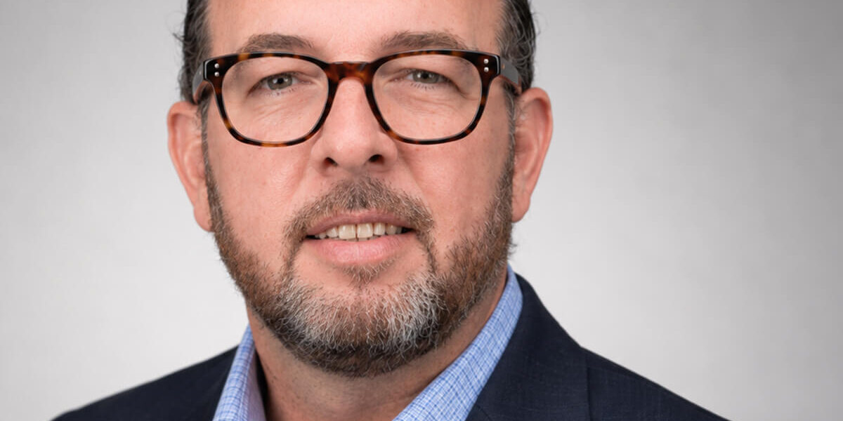 Professional headshot of a man wearing tortoiseshell glasses, a navy blazer, and a light blue shirt against a gray studio background.