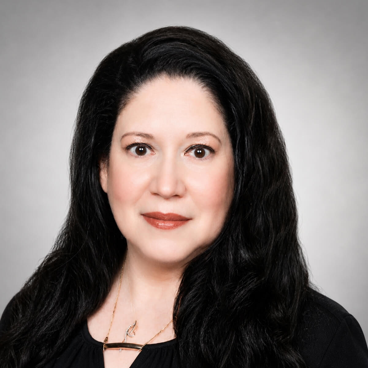 Head-and-shoulders portrait of a woman with long dark hair, wearing a black top and gold necklace, looking at the camera with a neutral expression.