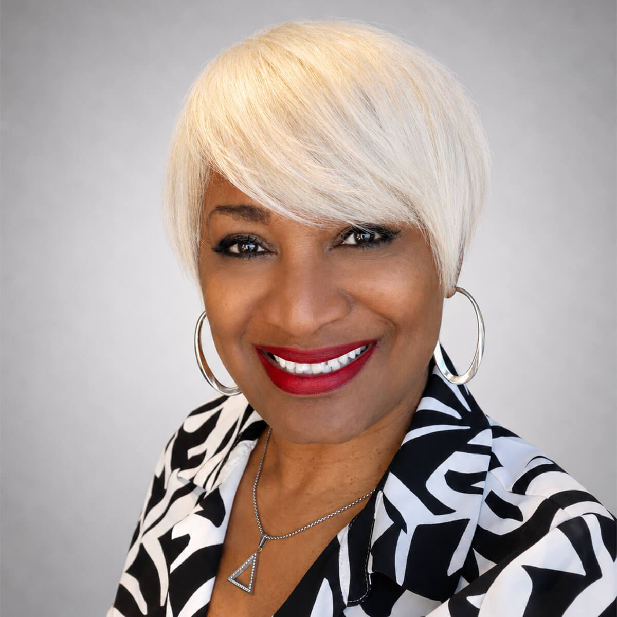 Smiling woman with short platinum blonde hair, hoop earrings, and a black-and-white patterned blouse portrait.