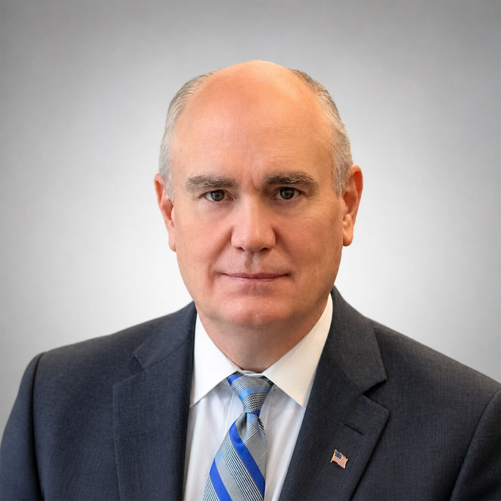 Professional head-and-shoulders portrait of a bald man in a dark suit and striped blue tie, wearing a small American flag pin on the lapel.