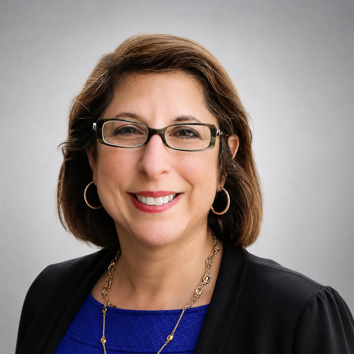 Professional headshot of a woman with short brown hair, glasses, gold hoop earrings, wearing a blue top and black blazer.