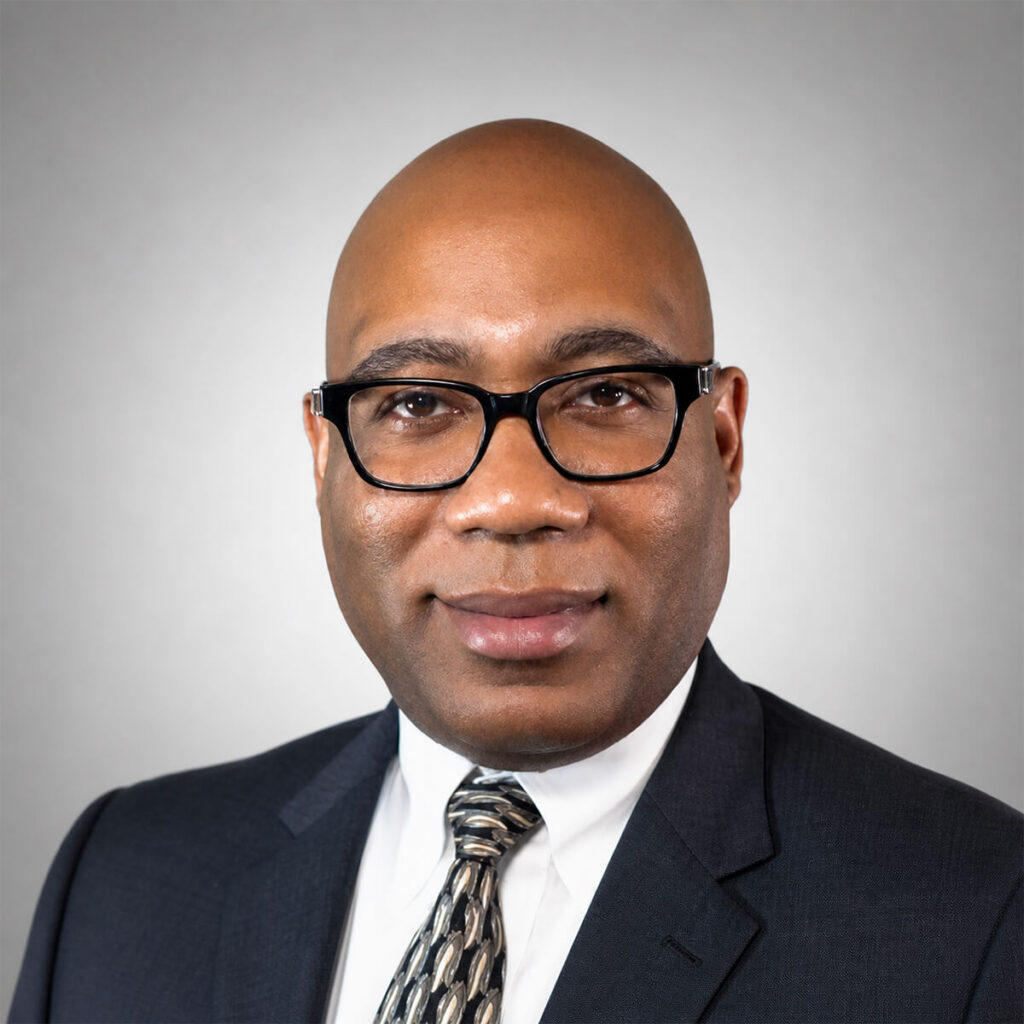 Headshot of a smiling, bald man wearing black-framed glasses, a white shirt, patterned tie, and a dark suit against a gray background.