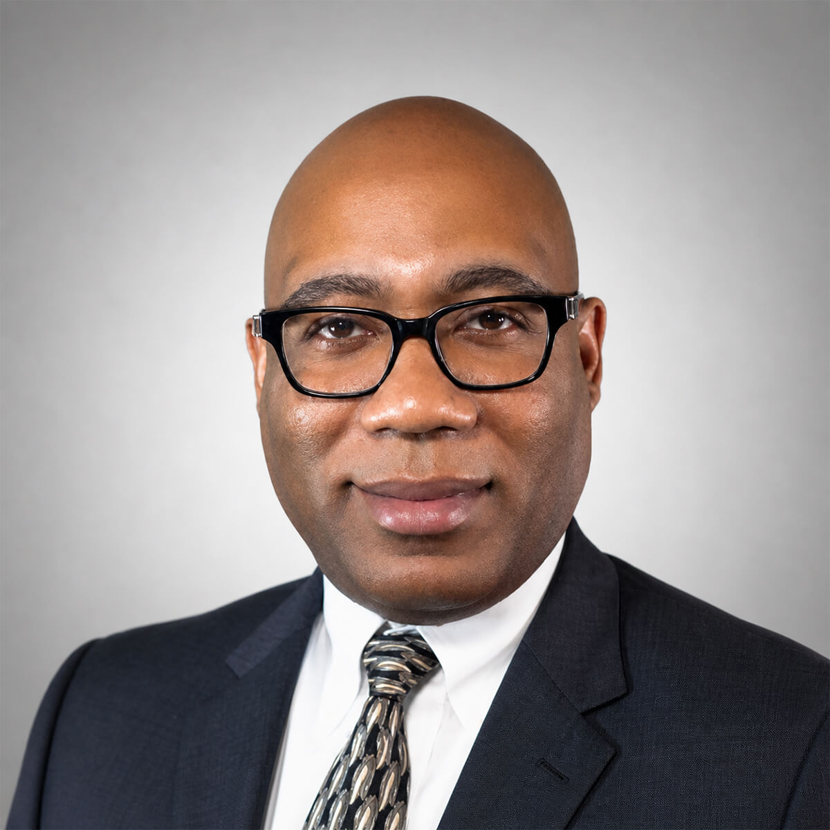 Headshot of a smiling, bald man wearing black-framed glasses, a white shirt, patterned tie, and a dark suit against a gray background.