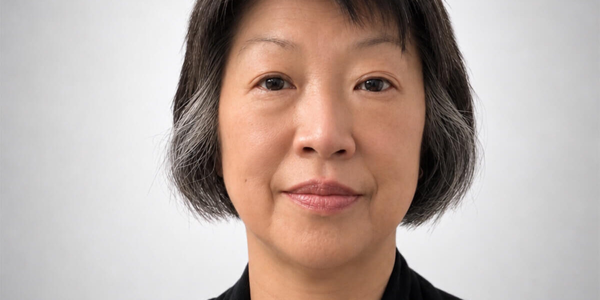 Yuen-Chun Close-up portrait of an Asian woman with short dark hair with gray strands, wearing a white lace top and black cardigan against a neutral gray background.