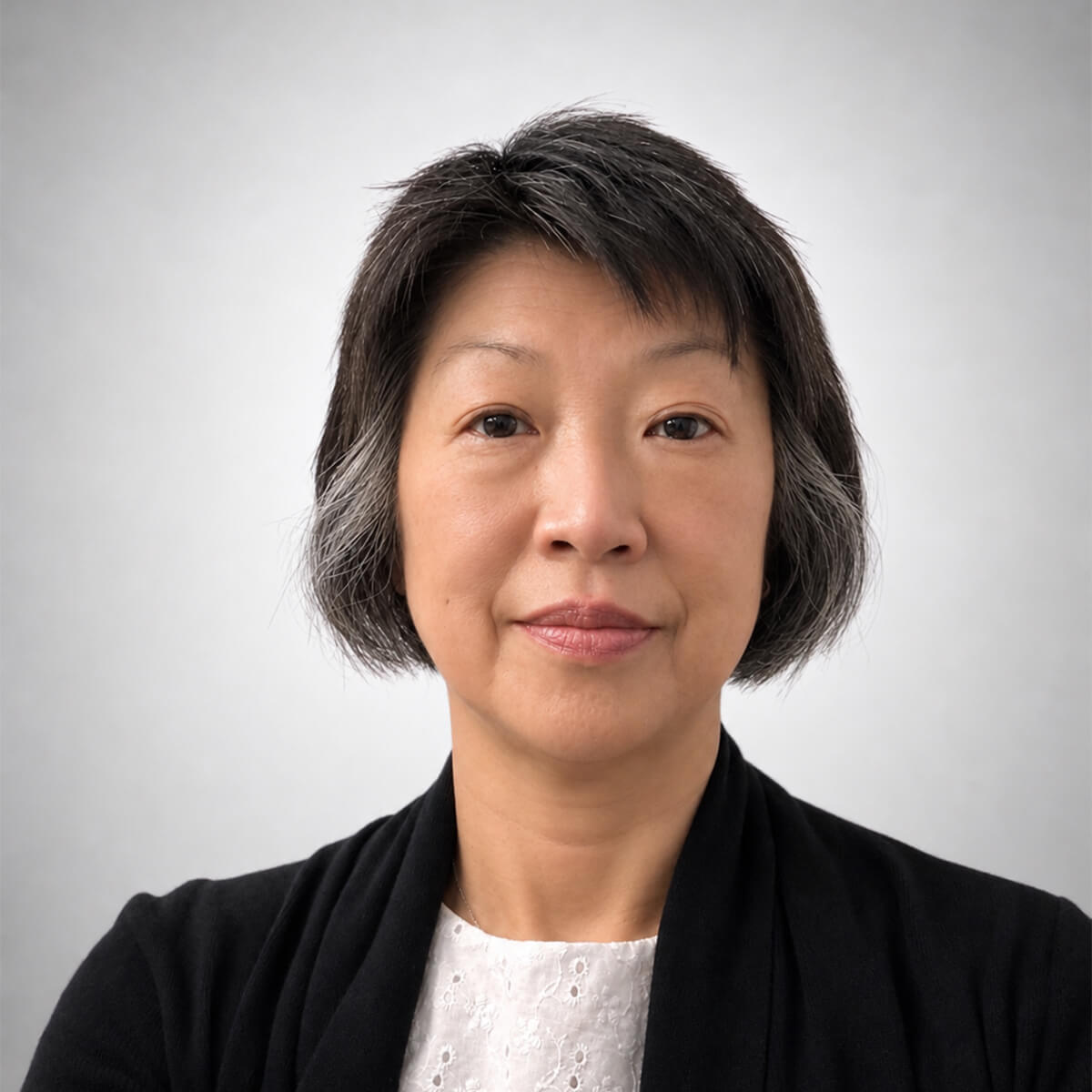 Close-up portrait of an Asian woman with short dark hair with gray strands, wearing a white lace top and black cardigan against a neutral gray background.