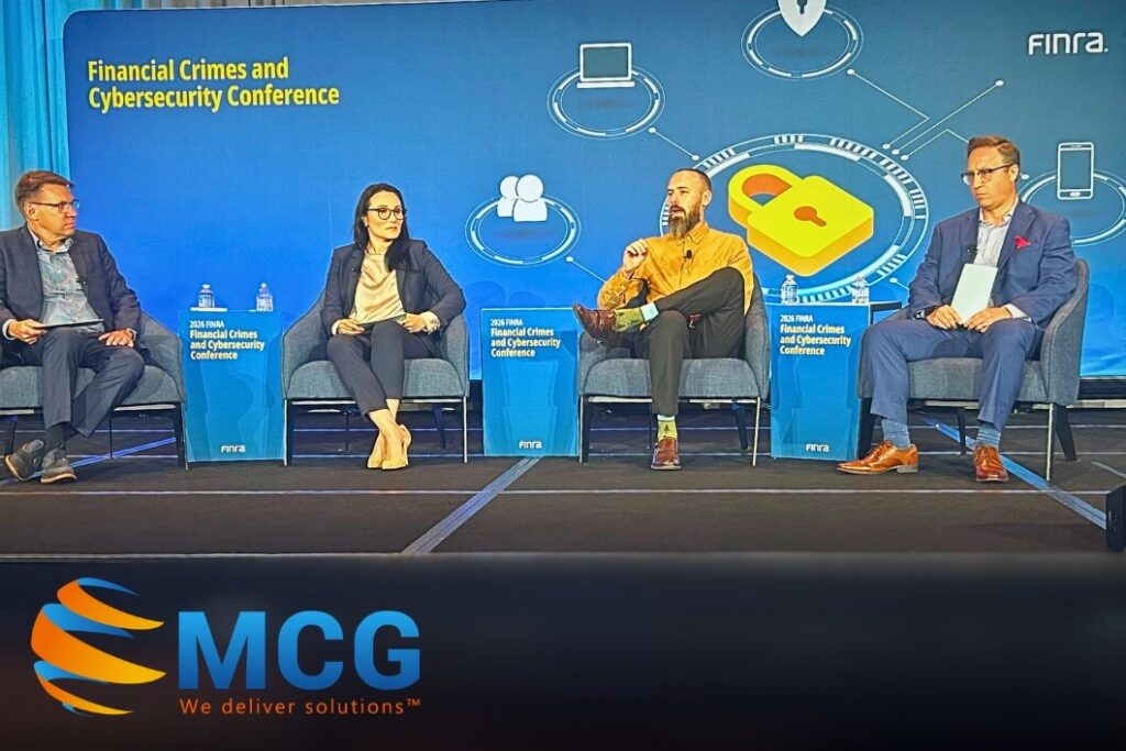 Panel discussion on stage at a Financial Crimes and Cybersecurity Conference with four speakers seated in blue chairs and a large blue backdrop featuring a lock icon and circuit graphics, MCG logo visible bottom left.